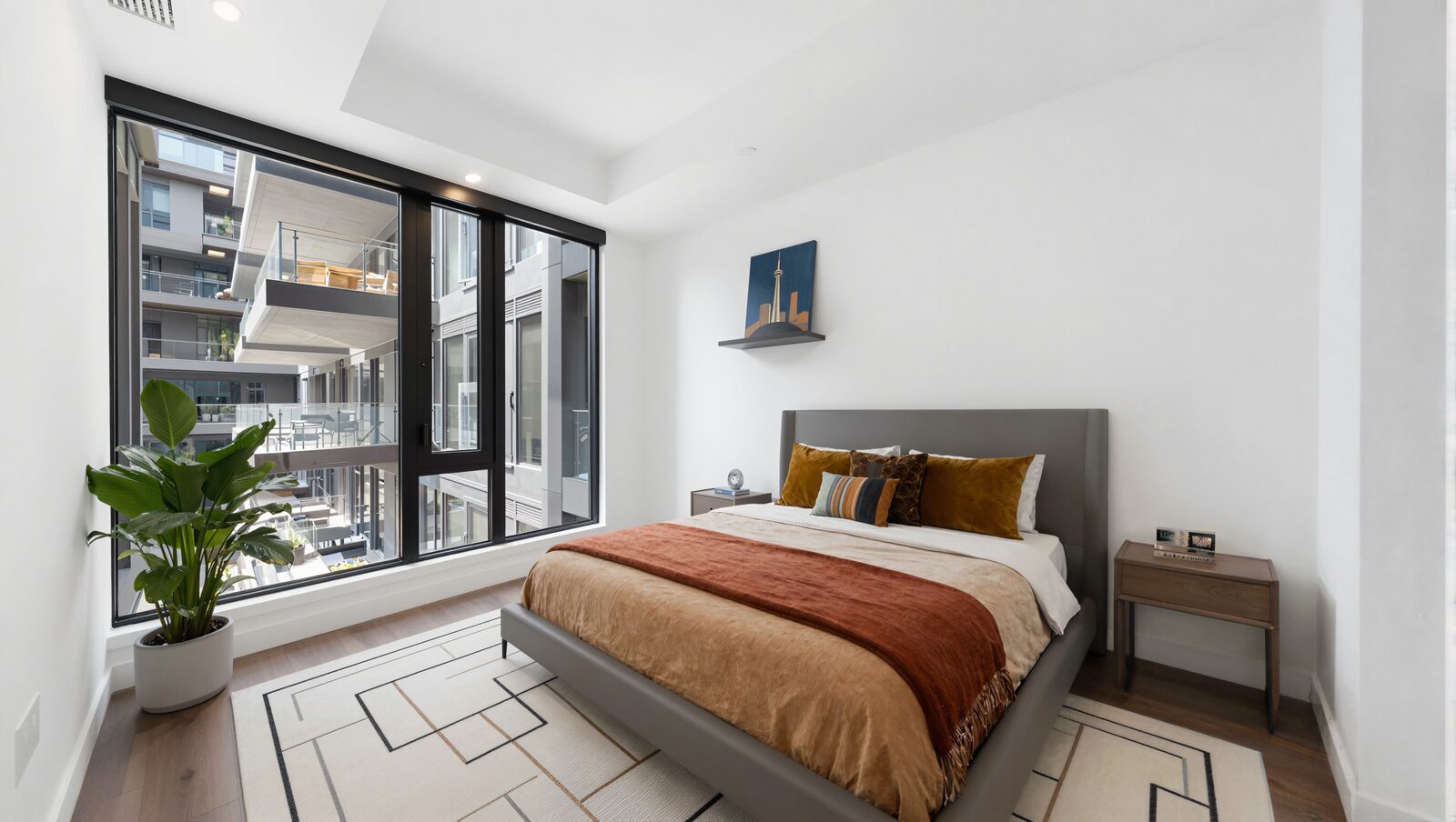 Primary bedroom virtually staged with a king bed, plant and Toronto skyline artwork beside a corner window
